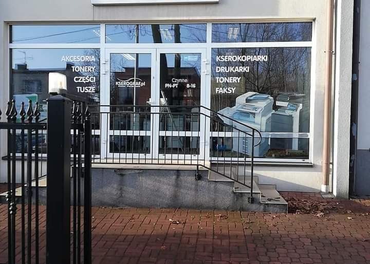 Fasada lokalu usługowego z dużymi witrynami, na których widnieją napisy: akcesoria, tonery, części, tusze, kserokopiarki, drukarki, tonery, faksy oraz godziny otwarcia, przed wejściem schody...