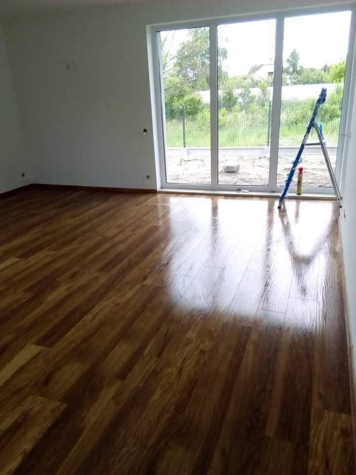 Świeżo umyta, lśniąca drewniana podłoga w pustym pokoju z dużym oknem, przez które widać ogród; przy oknie stoi niebieska drabina i butelka płynu do mycia.