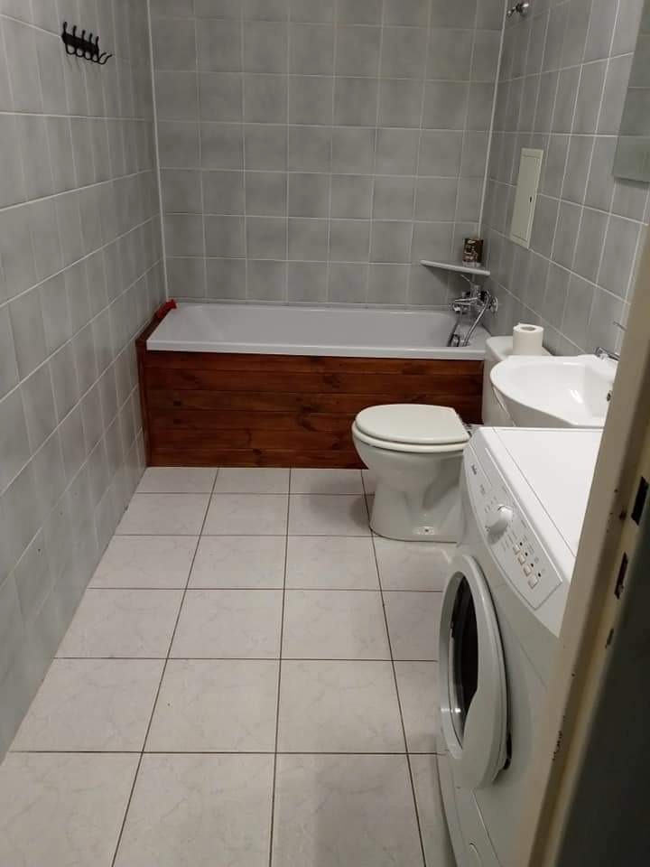 Wnętrze łazienki z wanną obudowaną ciemnym drewnem, białą toaletą, umywalką i pralką, wyłożonej szarymi i białymi płytkami.