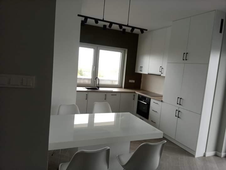 Jasna, nowocześnie urządzona kuchnia z białymi szafkami, drewnianym blatem i białym stołem z krzesłami, oświetlona czarną listwą z reflektorami.