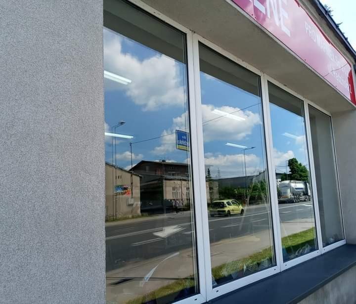 Duże, czyste okna wystawowe z białymi ramami, odbijające błękitne niebo z chmurami i uliczny ruch, widoczne budynki po drugiej stronie ulicy.