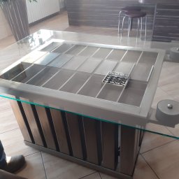 Łukasz - Stół z blatem ze szkła hartowanego, metalową konstrukcją z przegródkami i podstawą z pionowych, drewnopodobnych listew.