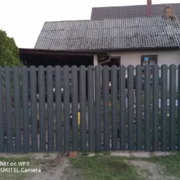 Nowoczesne ogrodzenie metalowe z pionowych profili w kolorze grafitowym, widoczny poziomica oparta o ogrodzenie, w tle budynek z szarą dachówką.