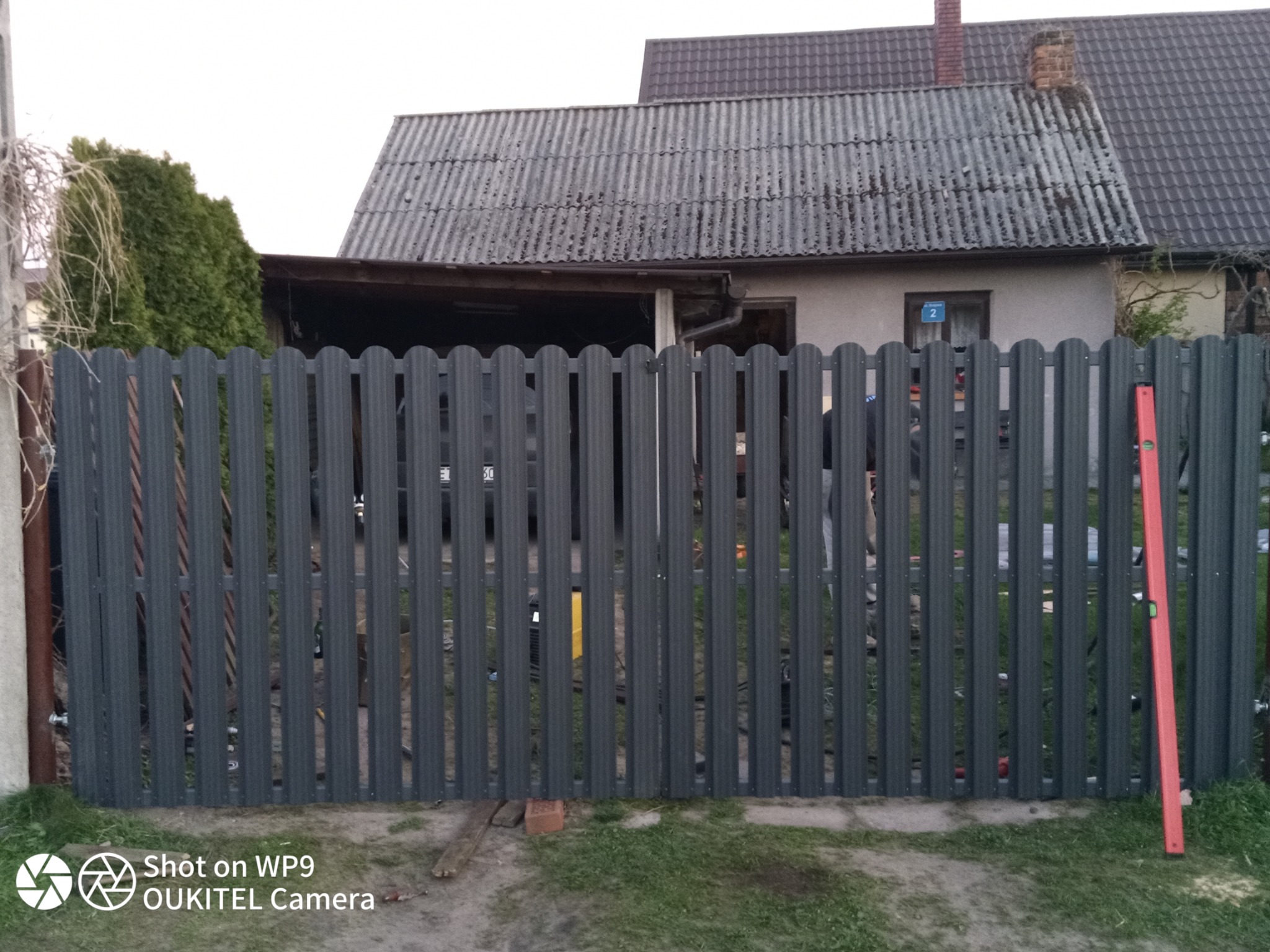 Nowoczesne ogrodzenie metalowe z pionowych profili w kolorze grafitowym, widoczny poziomica oparta o ogrodzenie, w tle budynek z szarą dachówką.