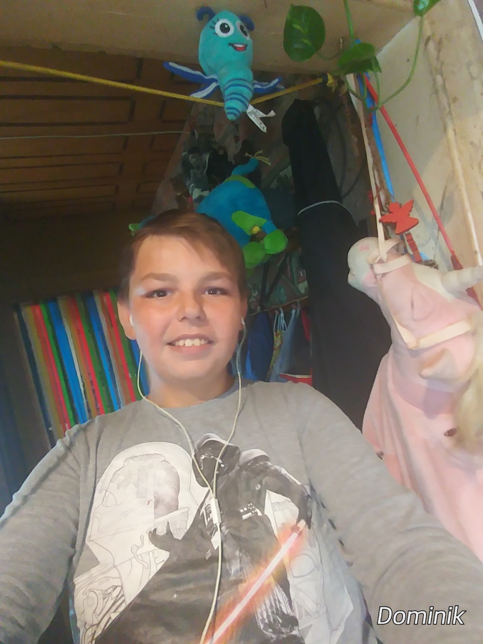 Uśmiechnięty chłopiec w szarej koszulce z motywem Star Wars, słuchający muzyki na słuchawkach, z pluszakami i dekoracjami w tle pokoju dziecięcego.