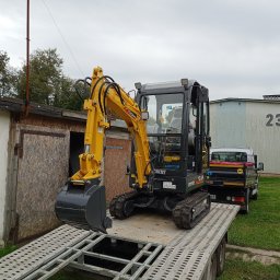 A.c.minikop Adam Ciostek - Żółto-czarna mini koparka Yuchai na platformie transportowej w Sokółce, z zaparkowanym samochodem dostawczym w tle i budynkiem z numerem 23.