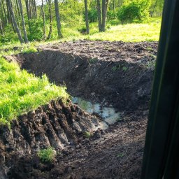 A.c.minikop Adam Ciostek - Świeżo wykopany rów wypełniony wodą, widok z kabiny maszyny, zielone drzewa w tle.