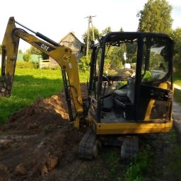 A.c.minikop Adam Ciostek - Żółta mini koparka CAT podczas prac ziemnych na działce, widoczne świeżo wykopane zagłębienie w ziemi, w tle wiejski krajobraz z drewnianym budynkiem.