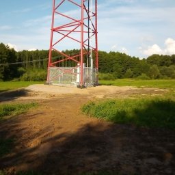 A.c.minikop Adam Ciostek - Stalowa wieża telekomunikacyjna pomalowana na biało i czerwono, wzniesiona na wyrównanym, piaszczystym terenie z widocznymi śladami prac ziemnych, otoczona zielenią drzew i trawą pod błękitnym...