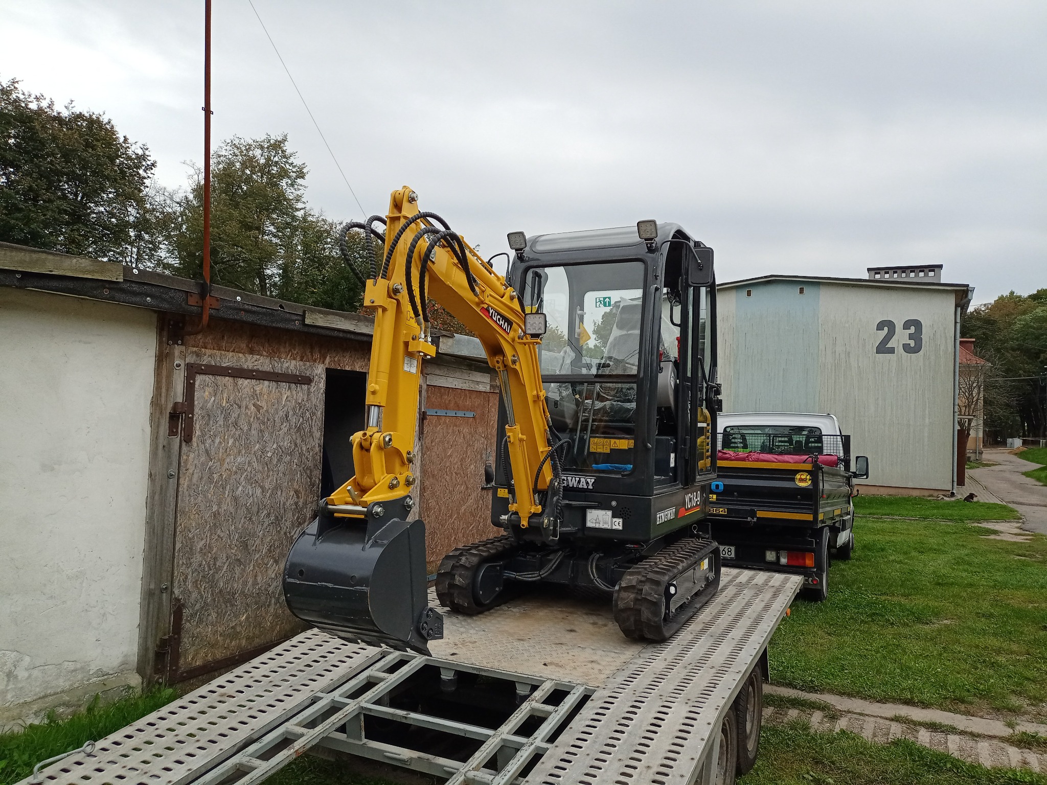 Żółto-czarna mini koparka Yuchai na platformie transportowej w Sokółce, z zaparkowanym samochodem dostawczym w tle i budynkiem z numerem 23.