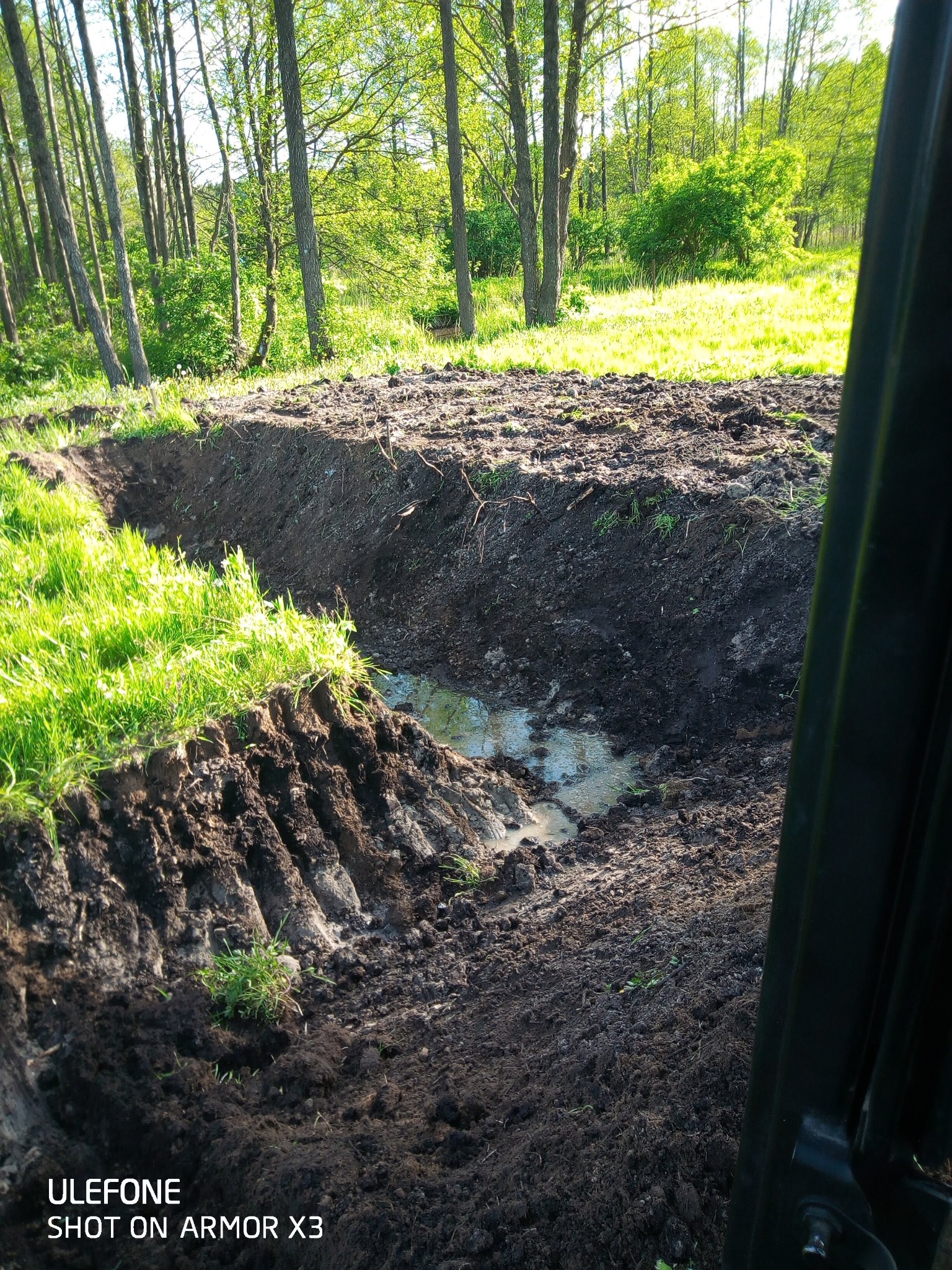 Świeżo wykopany rów wypełniony wodą, widok z kabiny maszyny, zielone drzewa w tle.