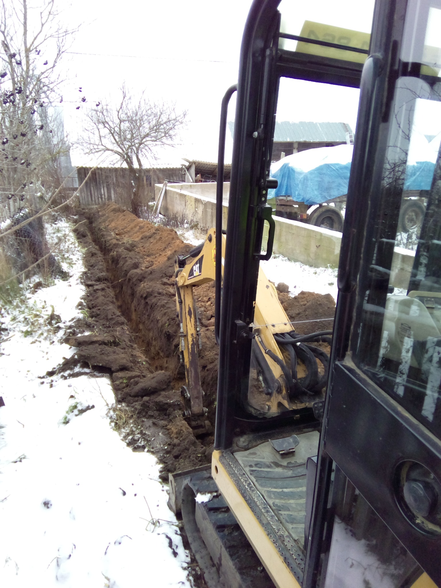 Żółta mini koparka CAT w trakcie wykopu wąskiego rowu w zimowej scenerii, widok z wnętrza kabiny operatora.