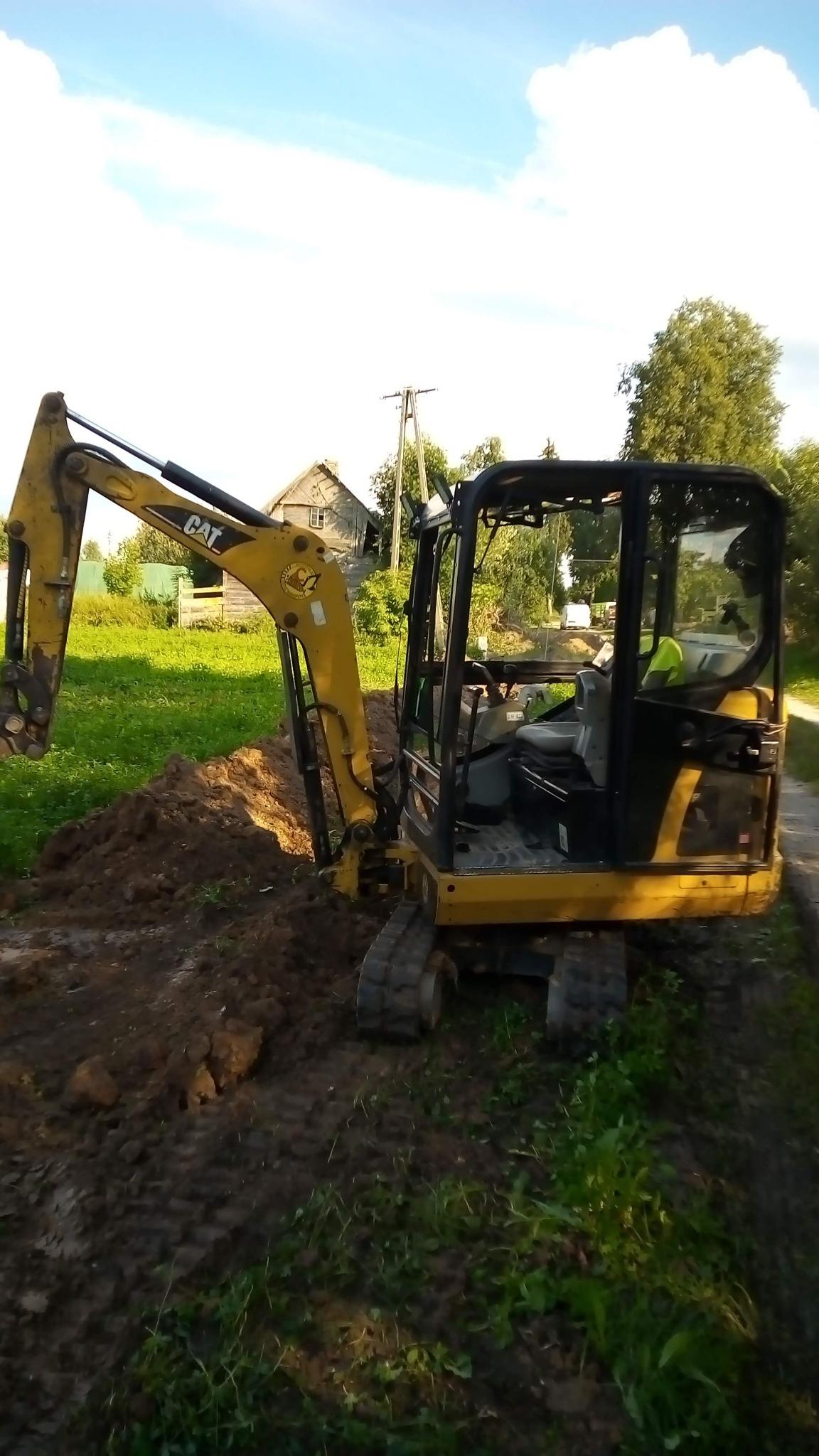 Żółta mini koparka CAT podczas prac ziemnych na działce, widoczne świeżo wykopane zagłębienie w ziemi, w tle wiejski krajobraz z drewnianym budynkiem.
