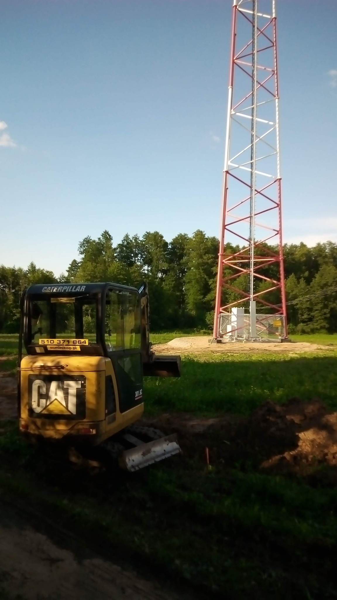 Żółta minikoparka Caterpillar 301.6C obok wykopu przy czerwono-białej wieży telekomunikacyjnej na tle zieleni i błękitnego nieba.
