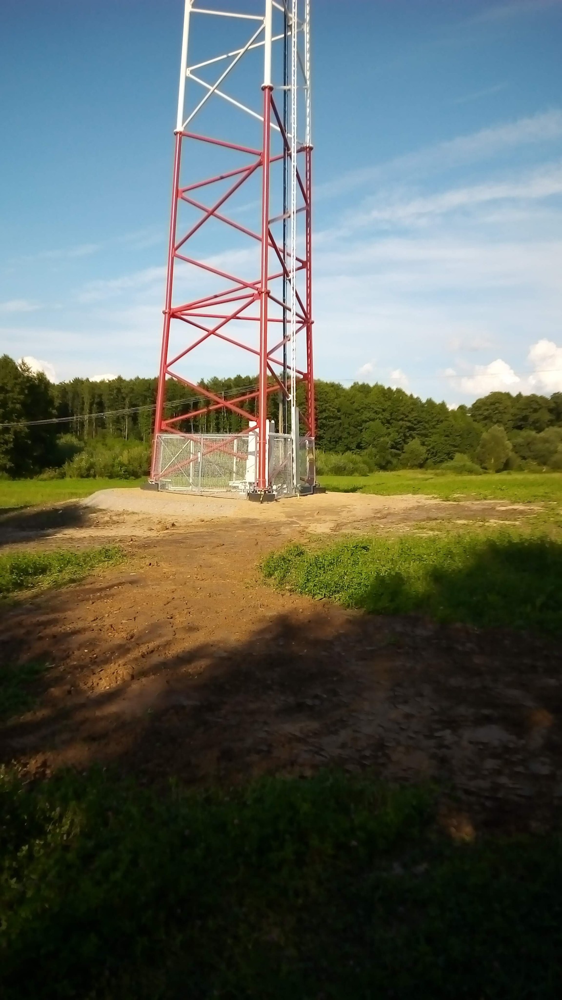 Stalowa wieża telekomunikacyjna pomalowana na biało i czerwono, wzniesiona na wyrównanym, piaszczystym terenie z widocznymi śladami prac ziemnych, otoczona zielenią drzew i trawą pod błękitnym...