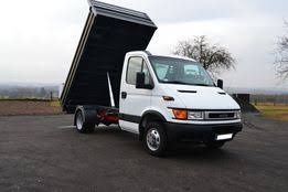 Biała wywrotka z podniesioną skrzynią ładunkową na asfaltowej powierzchni, widok z boku, w tle drzewa i pagórkowaty teren.