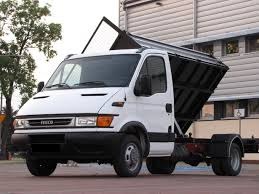 Biała wywrotka Iveco z podniesioną skrzynią ładunkową na tle budynku z szarymi panelami. Widoczne koła i podwozie pojazdu.