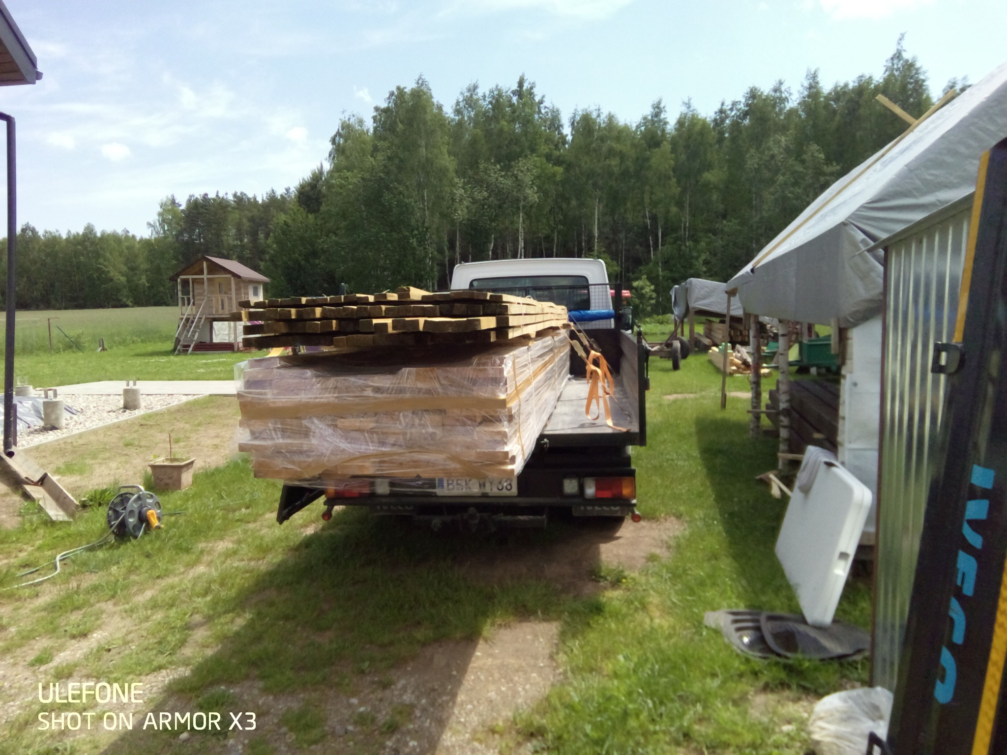 Ciężarówka Iveco z załadunkiem drewna konstrukcyjnego, zabezpieczonego folią, na tle zielonego trawnika, drzew i małego drewnianego domku.