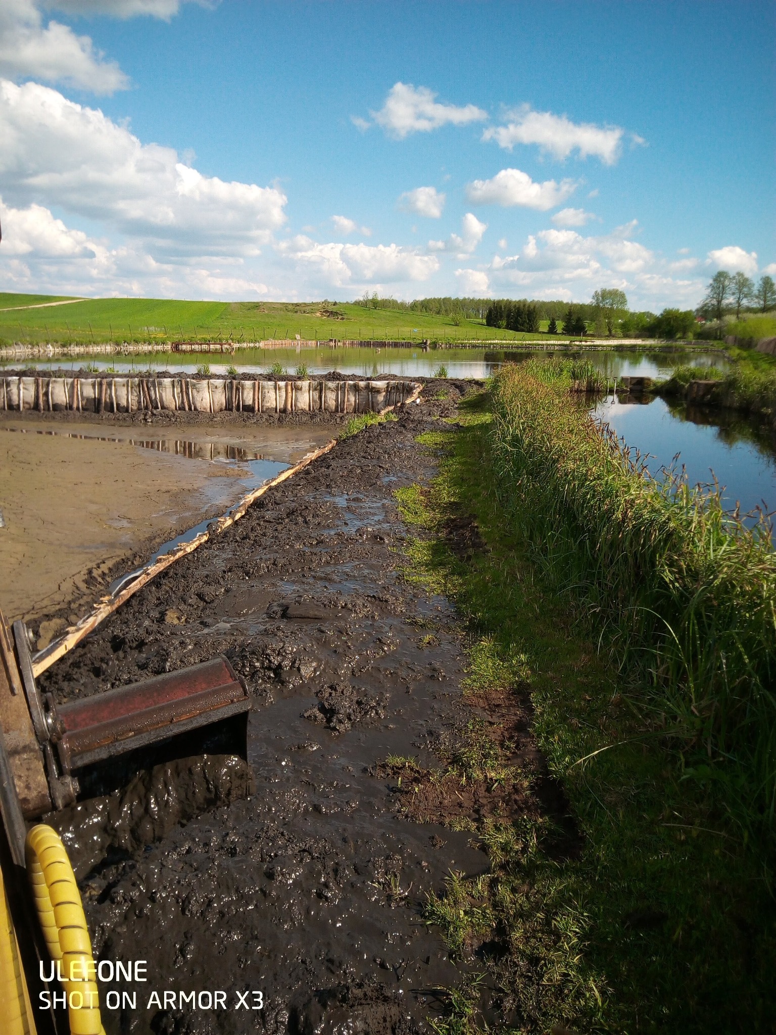 Częściowo osuszony staw z widocznym błotem, fragmentem maszyny i zielenią brzegów, pod błękitnym niebem z obłokami.