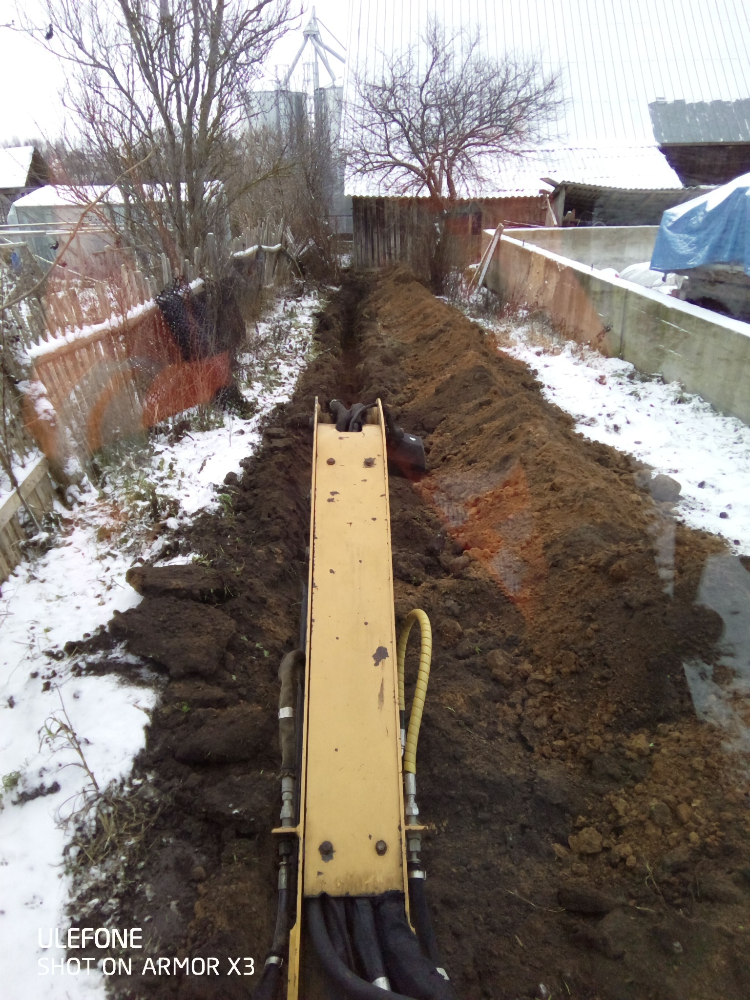 Wąski wykop w ziemi, częściowo pokrytej śniegiem, widziany z perspektywy żółtego ramienia koparki, w tle zabudowania gospodarcze i drzewa.