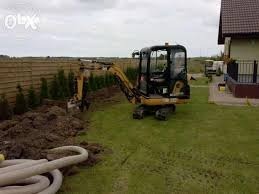 Mini koparka na trawniku obok wykopu i rur kanalizacyjnych, ogrodzenie panelowe w tle, pochmurne niebo.