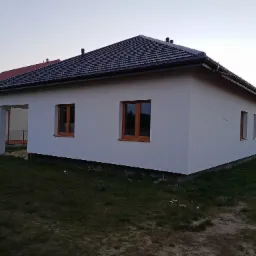 Nowo otynkowany, parterowy dom jednorodzinny z ciemną dachówką, widok z boku na tle zachodzącego słońca.