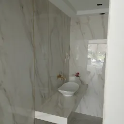 Nowoczesna łazienka w trakcie wykańczania z marmurowymi płytkami, złotą armaturą i białą umywalką nablatową, widoczne niedokończone prace przy suficie i surowa posadzka.