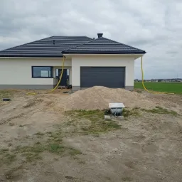 Nowo wybudowany, parterowy dom jednorodzinny z szarą bramą garażową i ciemną dachówką, otoczony placem budowy z piaskiem, paletami i żółtym wężem, na tle zielonego pola i pochmurnego nieba.