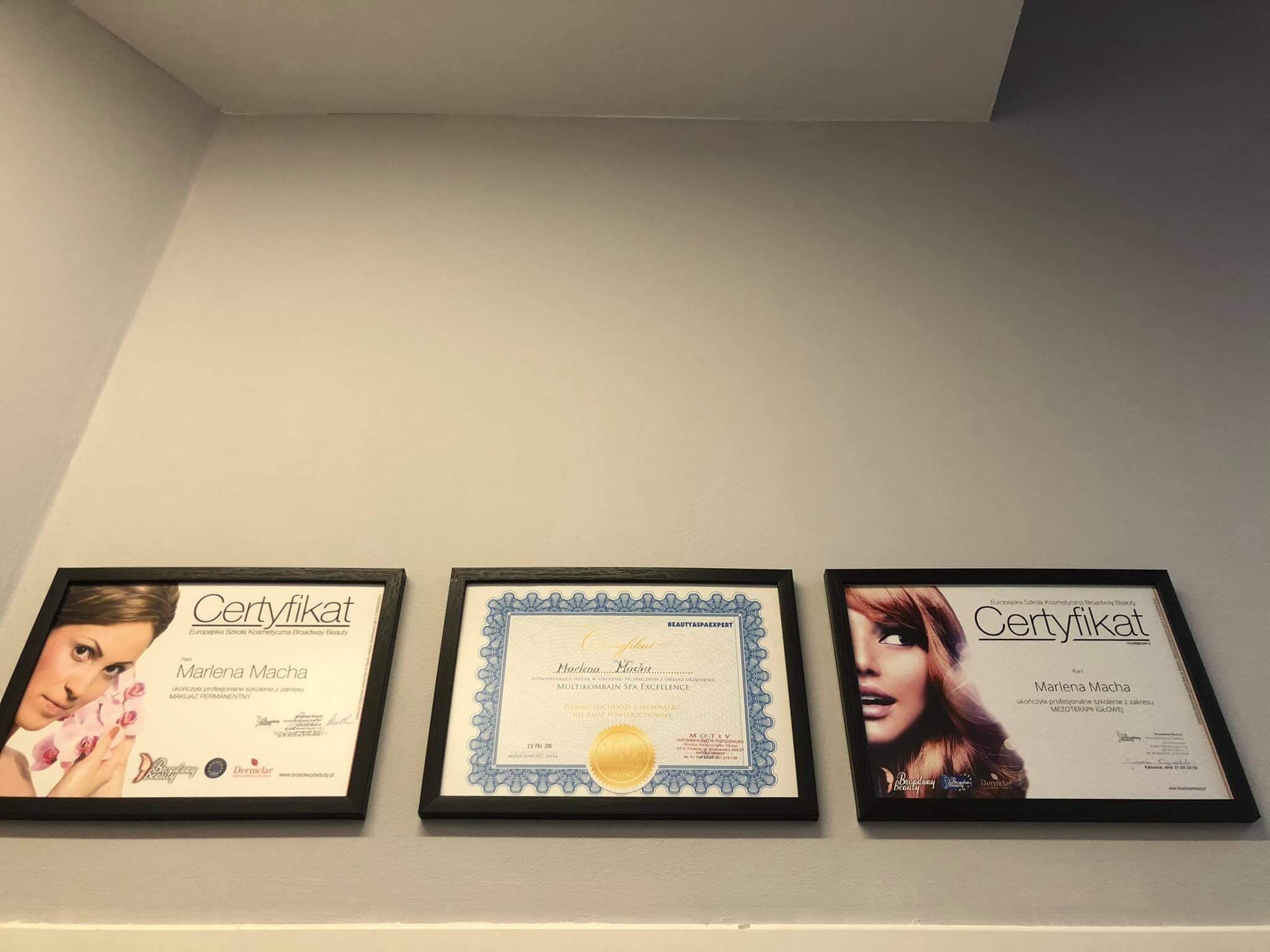 Trzy oprawione certyfikaty ukończenia szkoleń kosmetycznych wiszące na szarej ścianie, z widocznymi zdjęciami modelek i nazwami szkół kosmetycznych.