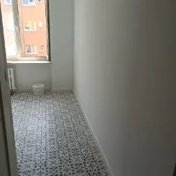 Pomalowane na biało wnętrze pokoju z wzorzystą, czarno-białą podłogą z płytek. Widoczne okno z widokiem na budynek mieszkalny i białe wiadro z farbą przy grzejniku.