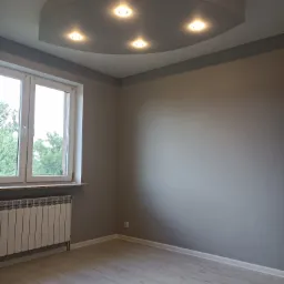 Pomalowane na szaro wnętrze pokoju z nową podłogą, grzejnikiem i oknem, w suficie owalna wnęka z pięcioma punktami świetlnymi.