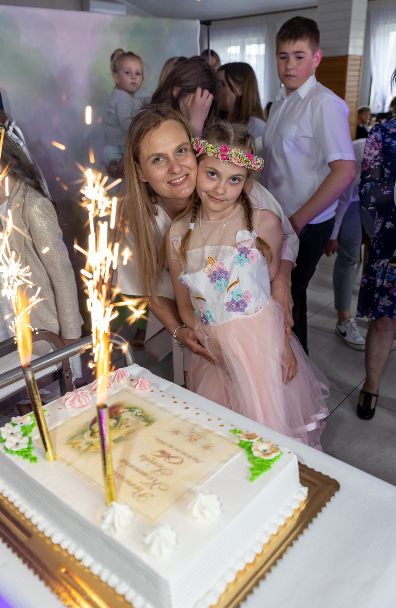 Uśmiechnięta mama obejmuje córkę w kwiecistym wianku, na pierwszym planie tort z płonącymi fontannami iskier, w tle goście.