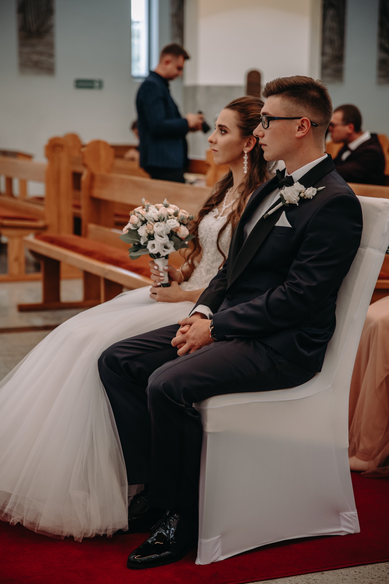 Para młoda w kościele, panna młoda trzyma bukiet, pan młody w okularach. W tle widoczni goście i fotograf.