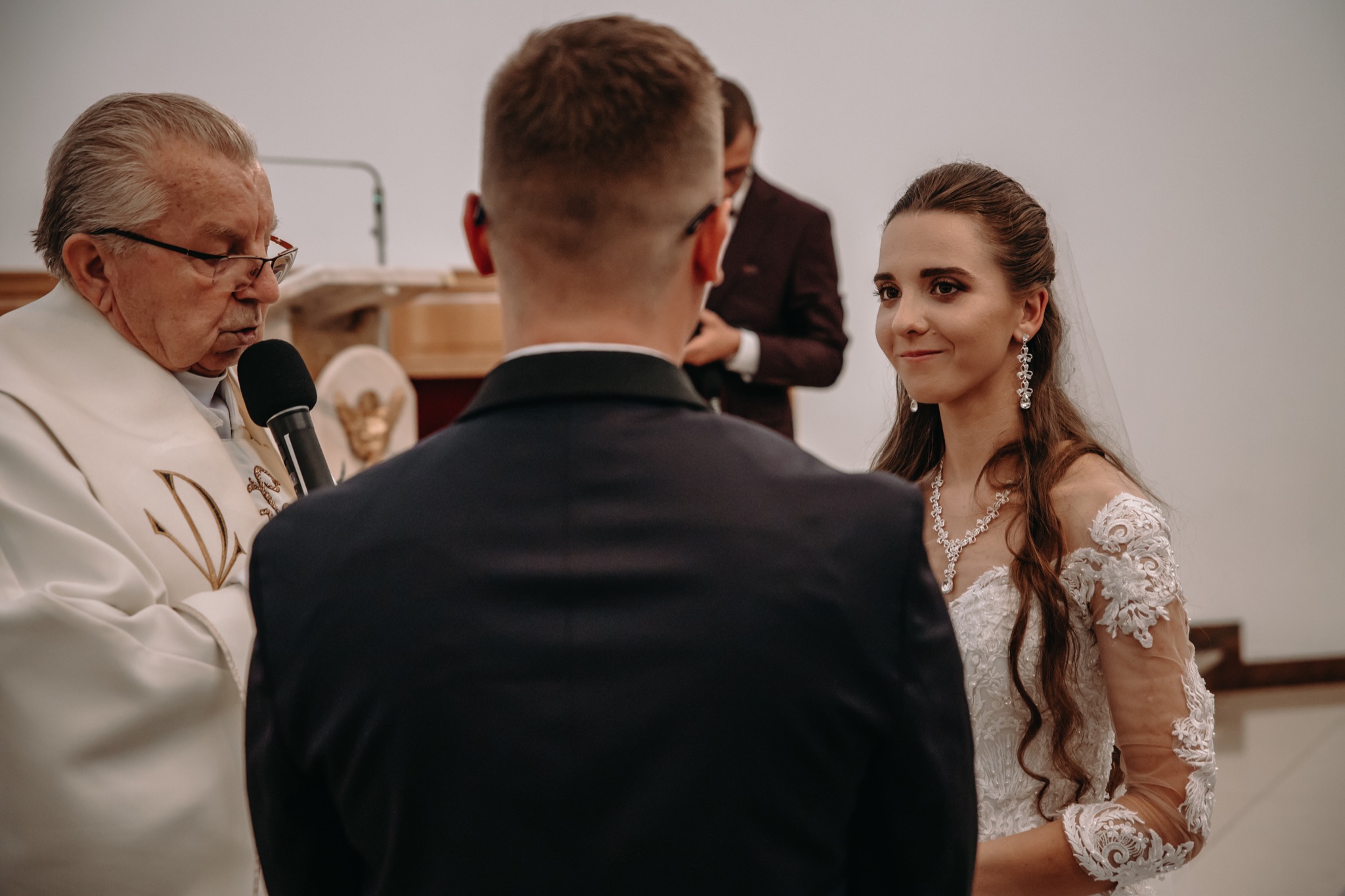 Panna młoda spogląda z uśmiechem na pana młodego podczas ceremonii ślubnej prowadzonej przez księdza w kościele.