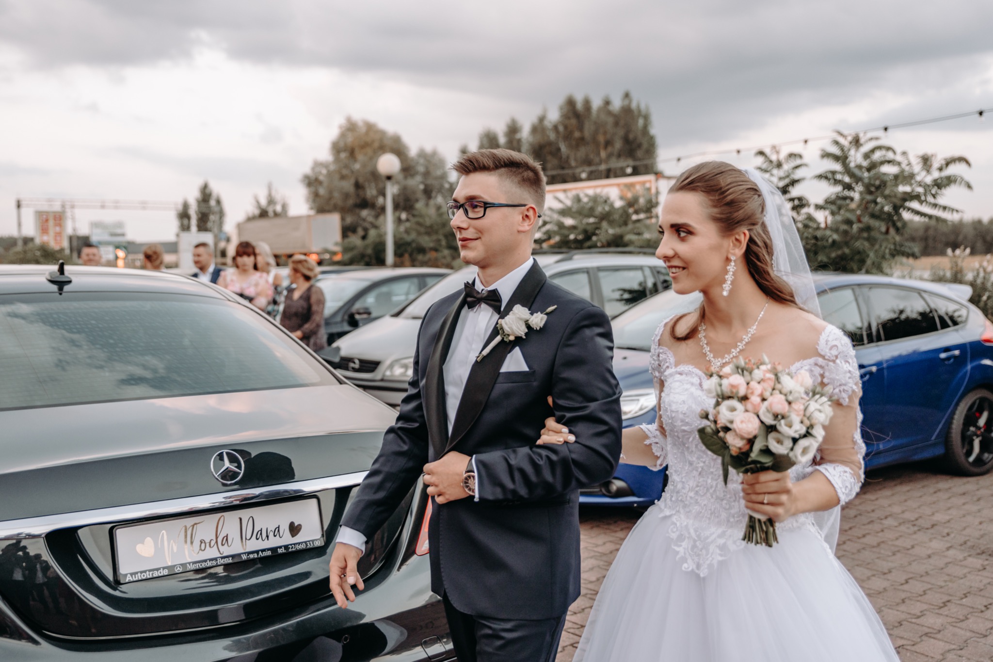 Para młoda w Warszawie, obok czarnego Mercedesa z tablicą 'Młoda Para', panna młoda w białej sukni z bukietem, pan młody w granatowym smokingu z muszką.