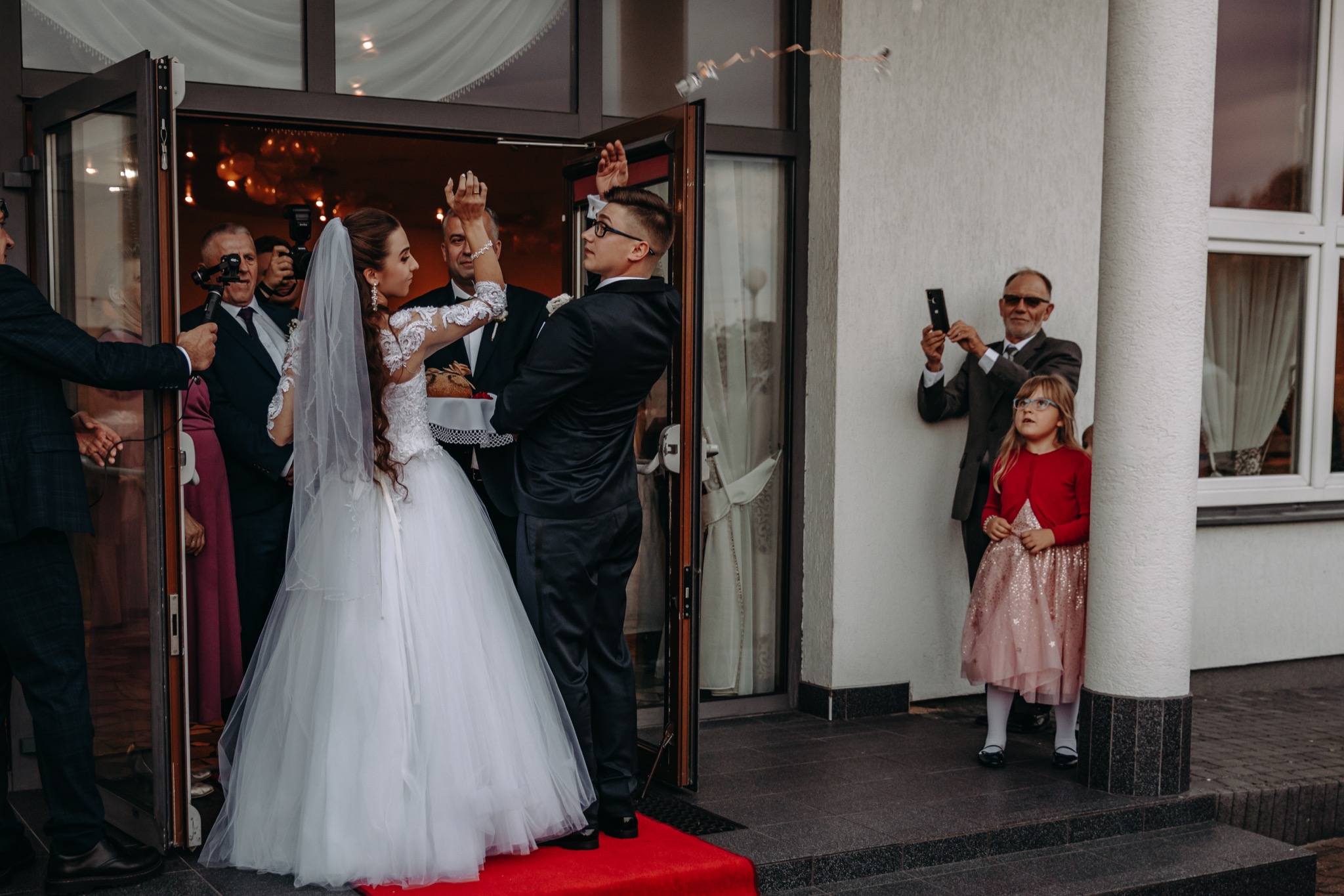 Para młoda wychodzi z budynku, obsypywana konfetti, goście robią zdjęcia telefonami, dziewczynka w różowej sukience obserwuje.
