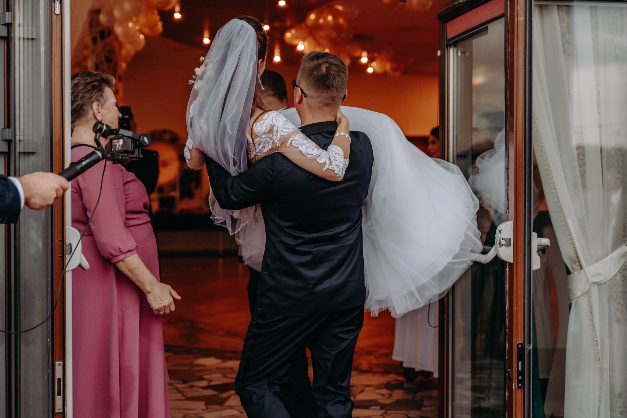 Pan młody w garniturze wnosi pannę młodą w sukni ślubnej przez drzwi, w tle goście i dekoracje balonowe, kobieta filmuje wydarzenie kamerą na stabilizatorze.