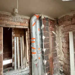 Świeżo otynkowane ściany z cegły w surowym wnętrzu, zabezpieczone rury owinięte folią i pomarańczową taśmą, widoczne listwy przypodłogowe i instalacja elektryczna.