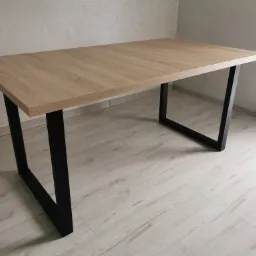 Stół z blatem o fakturze jasnego drewna i czarnymi, metalowymi nogami w kształcie prostokątów, ustawiony na jasnej podłodze w pomieszczeniu o jasnych ścianach.