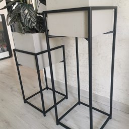 Global Welding - Dwie minimalistyczne, czarne, metalowe konstrukcje na donice z białymi, kwadratowymi wkładami, jedna z rośliną w tle ściana z delikatną fakturą.