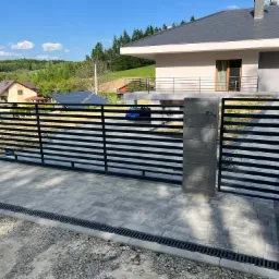 Nowoczesna, przesuwna brama wjazdowa z poziomych, czarnych profili, osadzona na szarych, betonowych słupkach, prowadząca na podjazd z kostki brukowej, z widocznym odpływem liniowym i fragmentem...