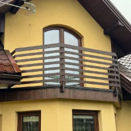 Żółty dom z brązowym dachem i balkonem z drewnianą balustradą o poziomym układzie szczebli, widoczne okno z łukiem nad balkonem i antena telewizyjna na dachu.
