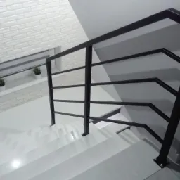 Nowoczesne schody z białą okładziną i czarną, geometryczną barierką z poziomych prętów, w tle ściana z białej cegły i okno z roślinami.