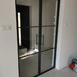 Dwuskrzydłowe drzwi wewnętrzne z czarnymi szprosami i przezroczystym szkłem, widoczne na tle białych ścian i marmurowej podłogi, z odkurzaczem w rogu.