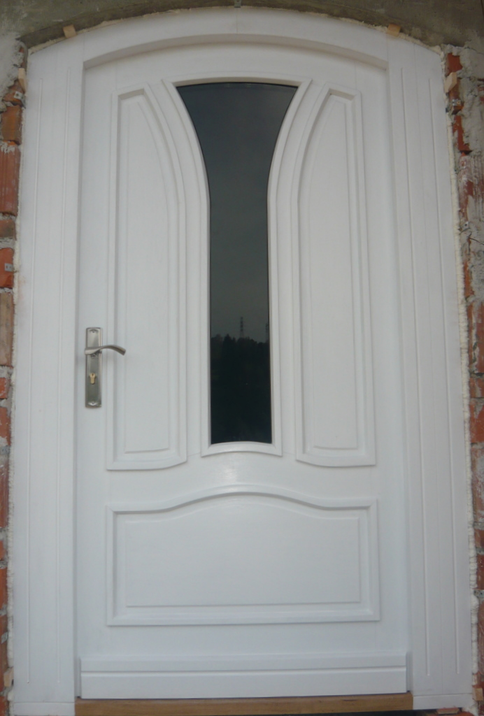 Białe drzwi wejściowe z szybą w kształcie łuku, widoczne ślady montażu na ścianie z cegły.