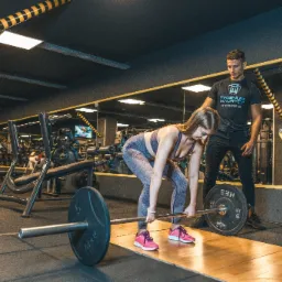 Kobieta w sportowym stroju wykonuje martwy ciąg ze sztangą w siłowni, pod nadzorem mężczyzny w koszulce z logo trenera.