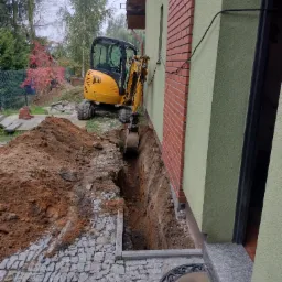 Żółta mini koparka JCB wykonuje wąski wykop wzdłuż ściany budynku z ceglaną wstawką, widoczny stos wykopanej ziemi i kostka brukowa.