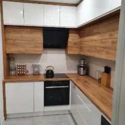 Nowoczesna kuchnia z białymi szafkami górnymi i dolnymi, drewnianymi blatami i panelami między szafkami, czarnym okapem i płytą indukcyjną, z widocznymi akcesoriami kuchennymi.