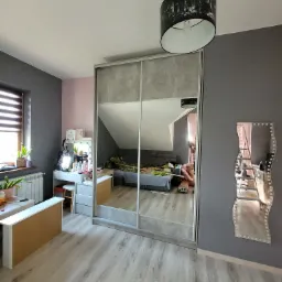 Duża szafa przesuwna z szarymi frontami lustrzanymi, odbijająca pokój na poddaszu z łóżkiem i osobą robiącą zdjęcie, obok toaletka z lustrem do makijażu i dekoracyjne lustro na ścianie