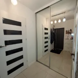 Przedpokój z dużą szafą przesuwną z lustrzanymi drzwiami, odbijającą wnętrze mieszkania z widoczną białą komodą i wieszakami na ubrania.